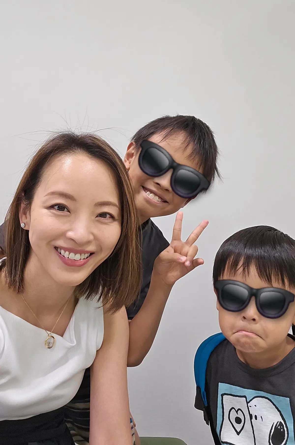 笑顔の女性と2人の男の子。女性は白いトップスにネックレスをつけ、男の子たちは黒いサングラスをかけている。1人はピースサイン、もう1人は少し不機嫌そうな表情。背景は白い壁。