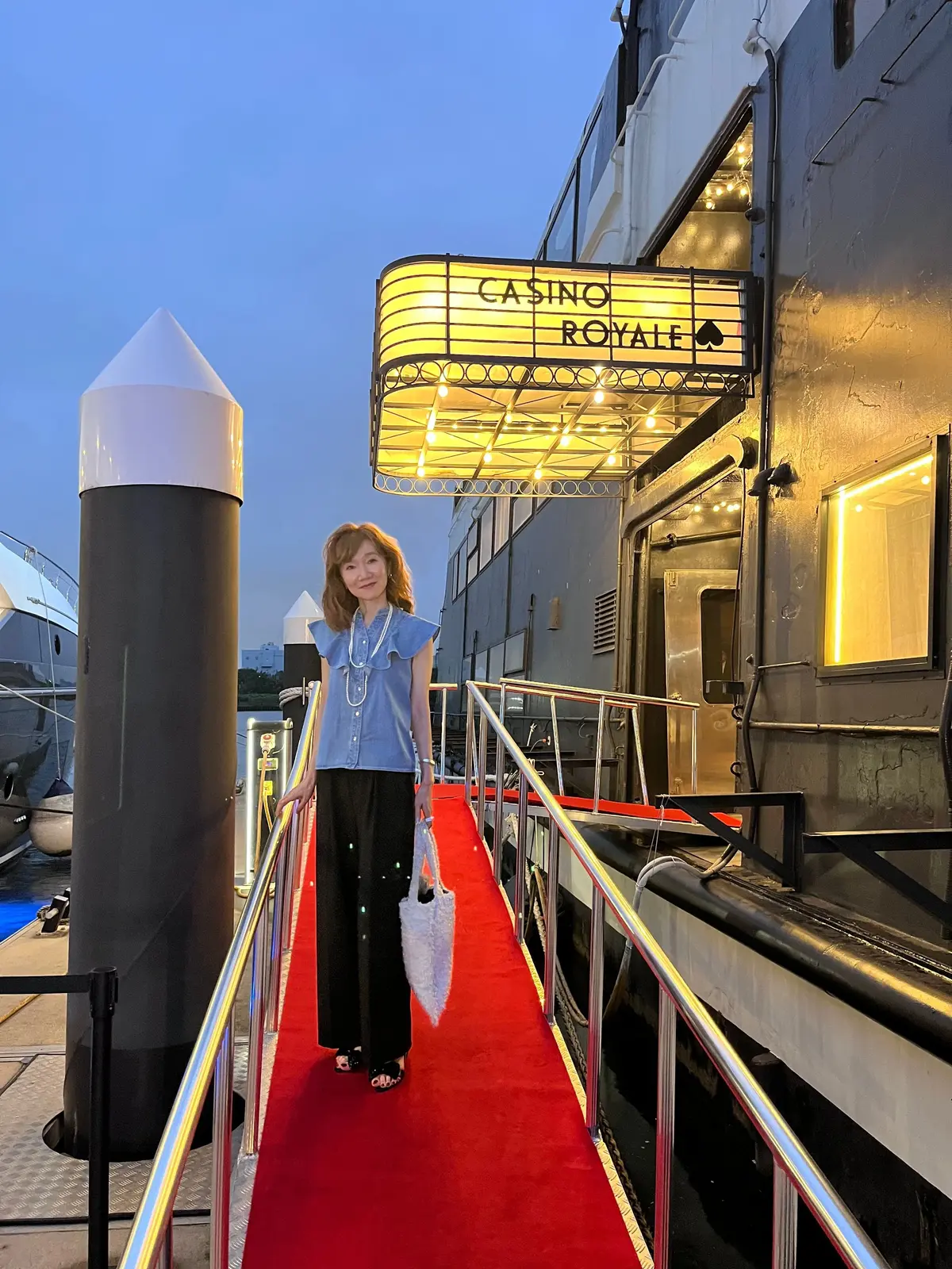 赤いカーペットが敷かれた通路に立つ女性。背景には「CASINO ROYALE」と光る看板のある船が見える。女性は青いシャツに黒いパンツを着用し、右手に赤いハンドバッグを持っている。空は夕焼けで、淡い青色。
