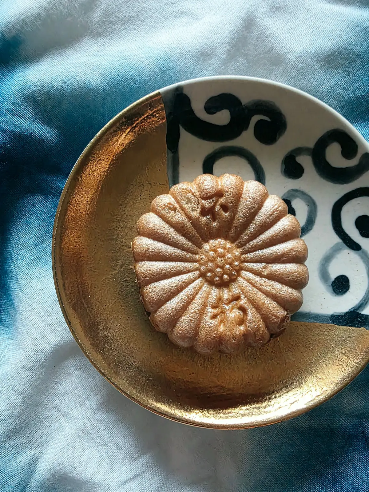 金色の浅いお皿に置かれた、菊の花のような模様の焼き菓子。表面は粉砂糖で覆われ、淡い茶色をしている。背景は青色の布。