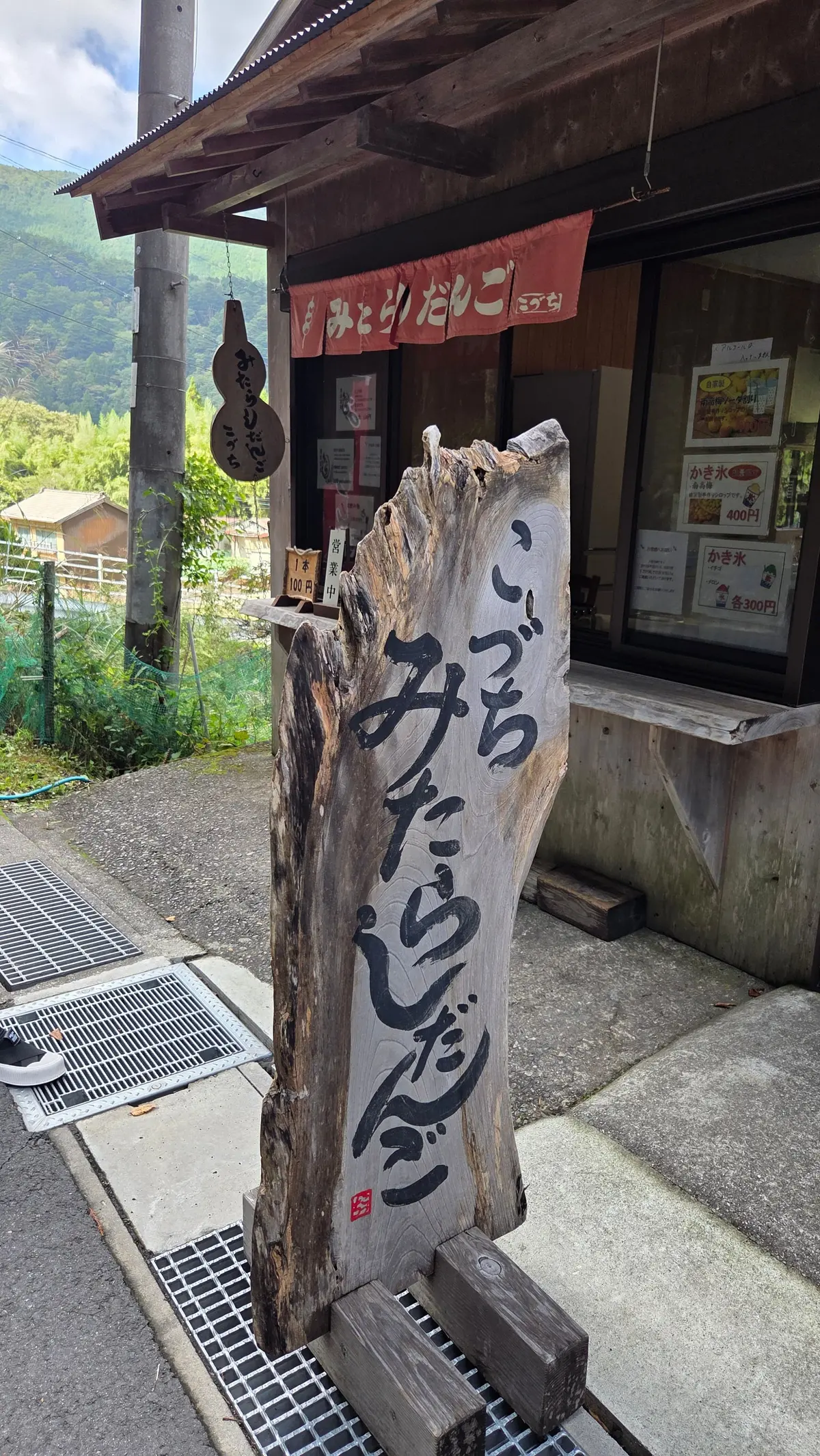木製の柱に墨で書かれた看板と、その奥にある小さな建物の写真。看板には「みやたのじゅうご」と大きく書かれ、建物の前には料金表が掲示されている。背景には緑豊かな山々が広がる。