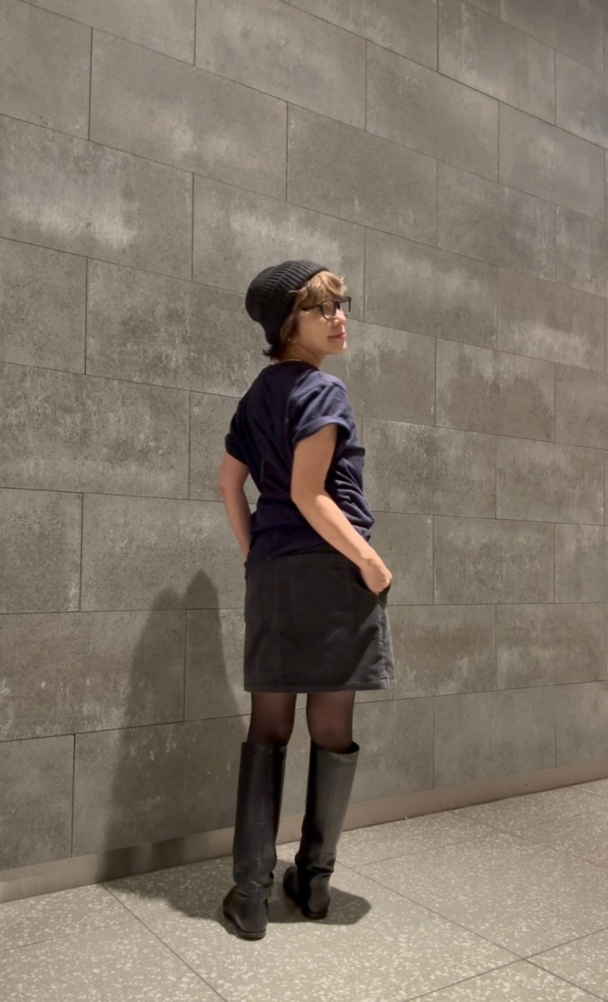 黒いニット帽を被った女性が、グレーのタイル壁を背に立っている。濃い青のTシャツ、黒のミニスカート、黒のタイツ、黒いロングブーツを着用。右手を腰に当て、こちらを振り返っている。