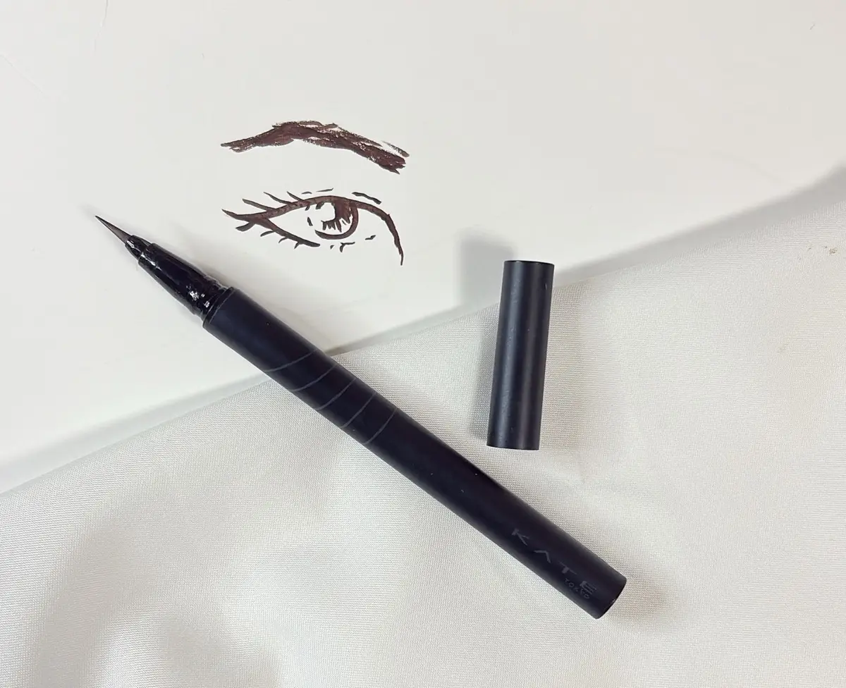 50代　40代　おすすめ　プチプラコスメ　KATE　ケイト　黒いアイライナーのペンと、描かれたアイラインのイラスト。ペンはマットブラックで、キャップが横に置かれている。白い布の上に置かれ、シンプルな構図。