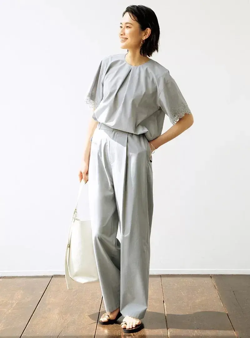 グレーのゆったりしたワンピースを着た女性が、木製の床の上に立ち、白いトートバッグを左手に持ち、笑顔でカメラを見つめている