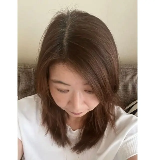 茶色の肩まである髪の女性が、白のTシャツを着てこちらを見下ろしている。顔は真ん中を捉えられており、やや影になっている。背景には茶色のソファがある。