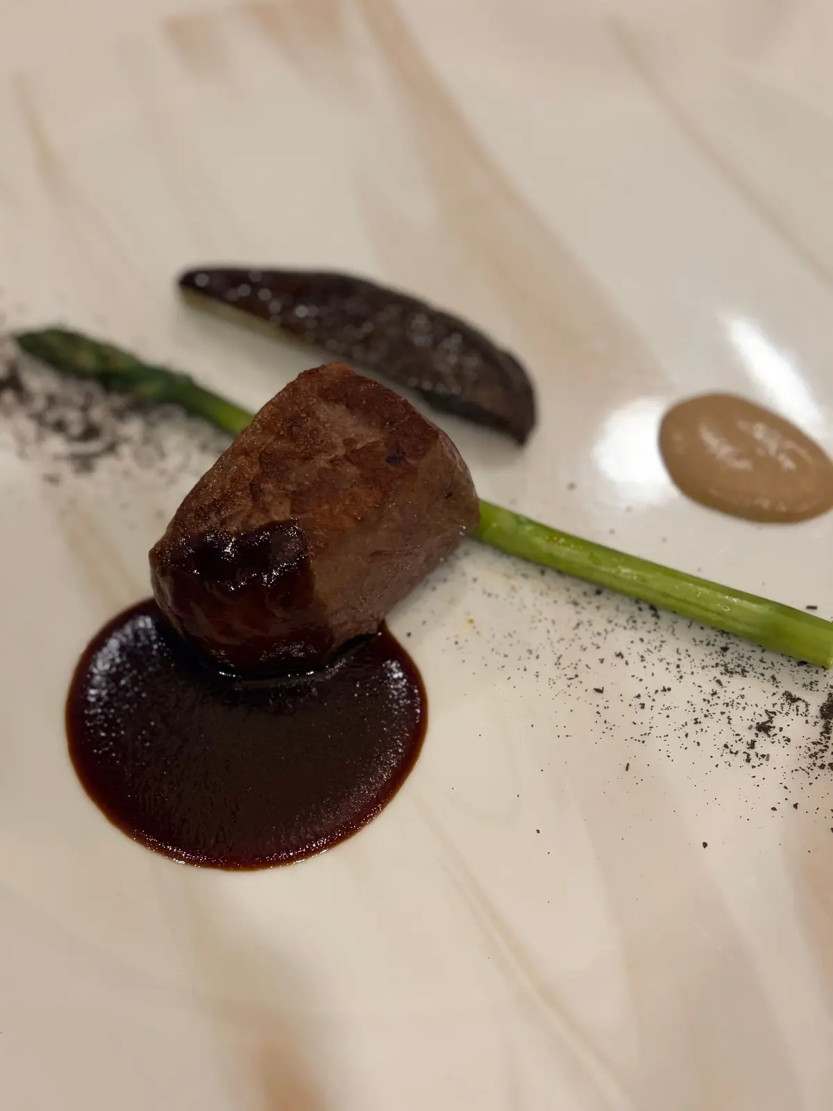 白磁の皿に盛り付けられた、牛肉の塊とソース、野菜の飾り。牛肉は濃い茶色で、円形の濃いソースが添えられている。皿の縁には、緑色の細長い野菜と、茶色のソースの点々、黒い粉末が散らばっている。