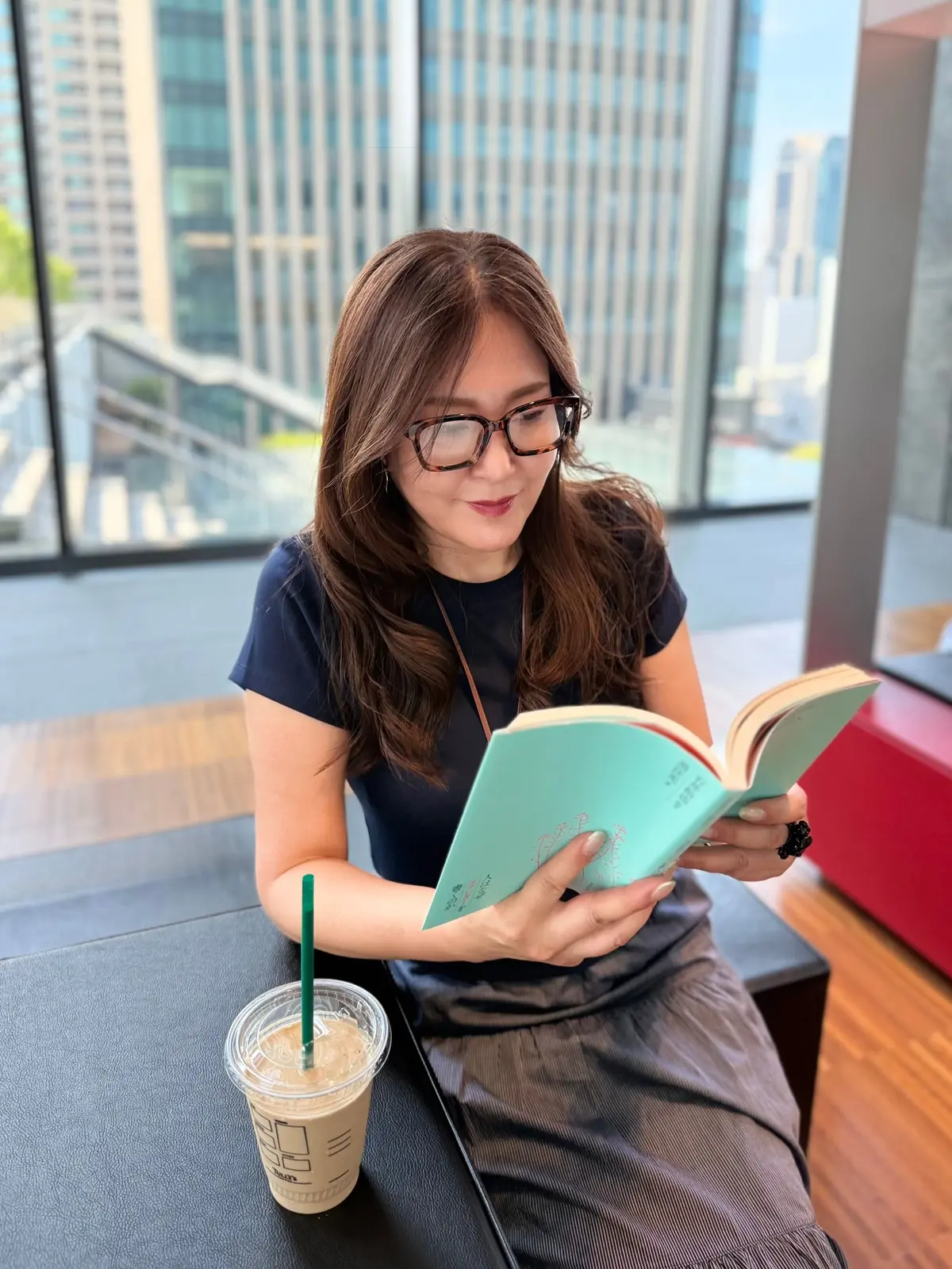 長髪の女性がミントグリーンの本を読み、窓際のテーブルに座っている。テーブルの上に緑のストローを入れた紙カップが置かれている。背景には高層ビルが並ぶ都市景観が見える。