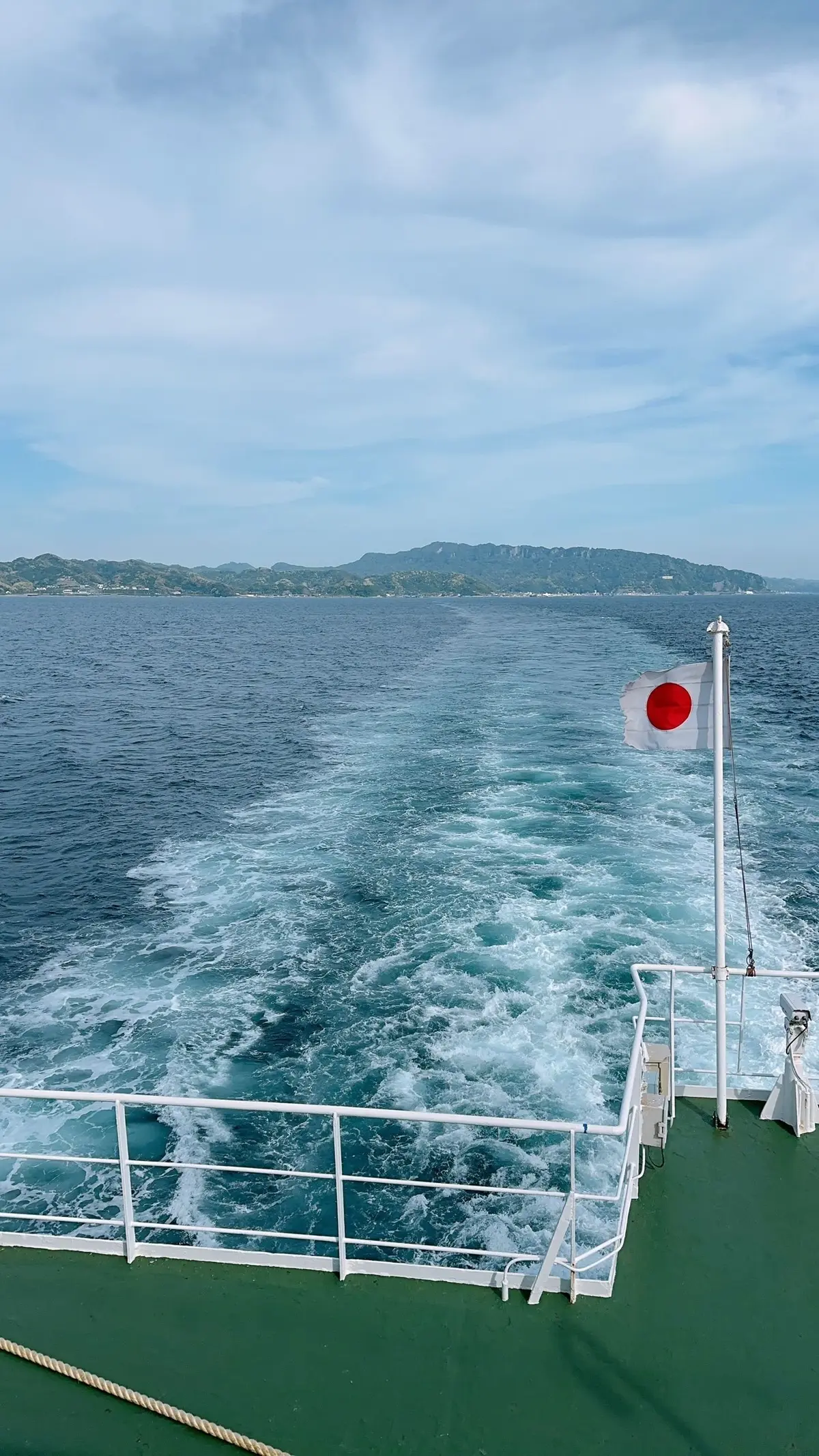 緑色の甲板から、水面を疾走する船の航跡と、その背後に見える緑豊かな海岸線。右には日本の国旗がはためいている。