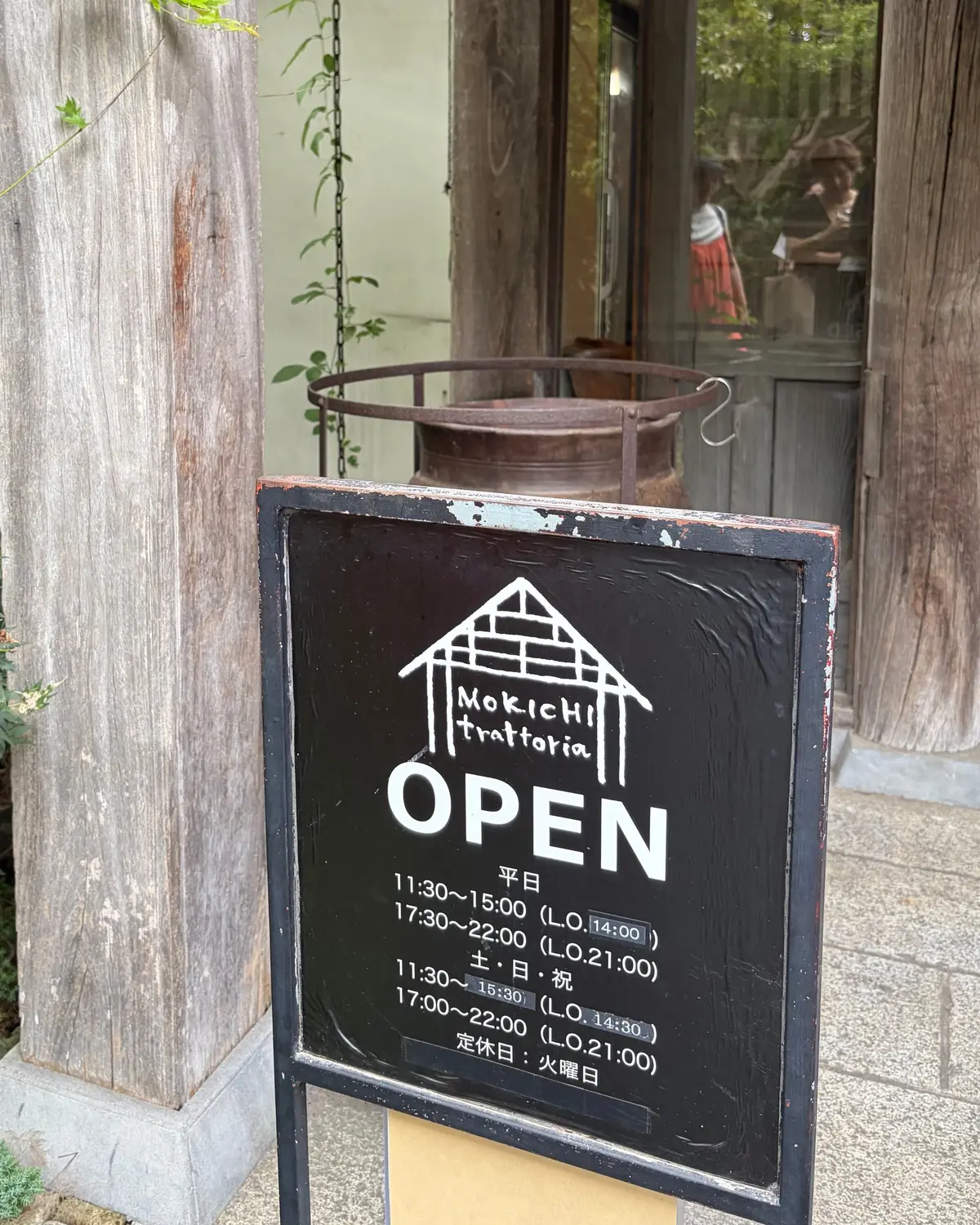 木目調の建物の入り口に設置された黒板の営業時間表示。上部に屋根のイラストと「Mokichi trattoria」の文字、中央に「OPEN」の文字がある。平日と土日祝日の営業時間がそれぞれ記載され、定休日も示されている。