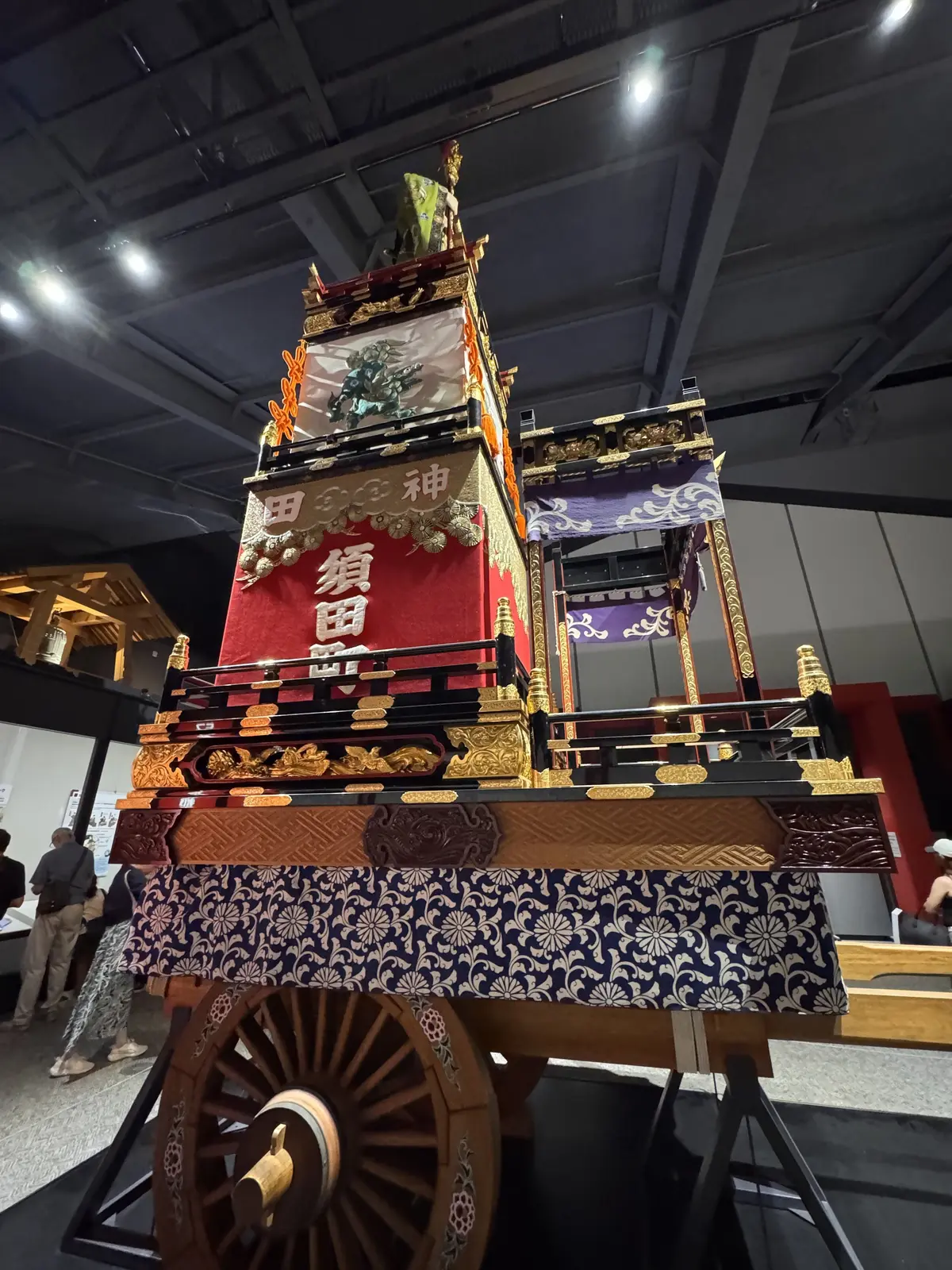 精巧に装飾された日本の山車。木製の台座に金色の装飾が施され、青い花柄の布で覆われている。最上部には神像が設置され、中央には日本語の文字が書かれた赤い幕が飾られている。