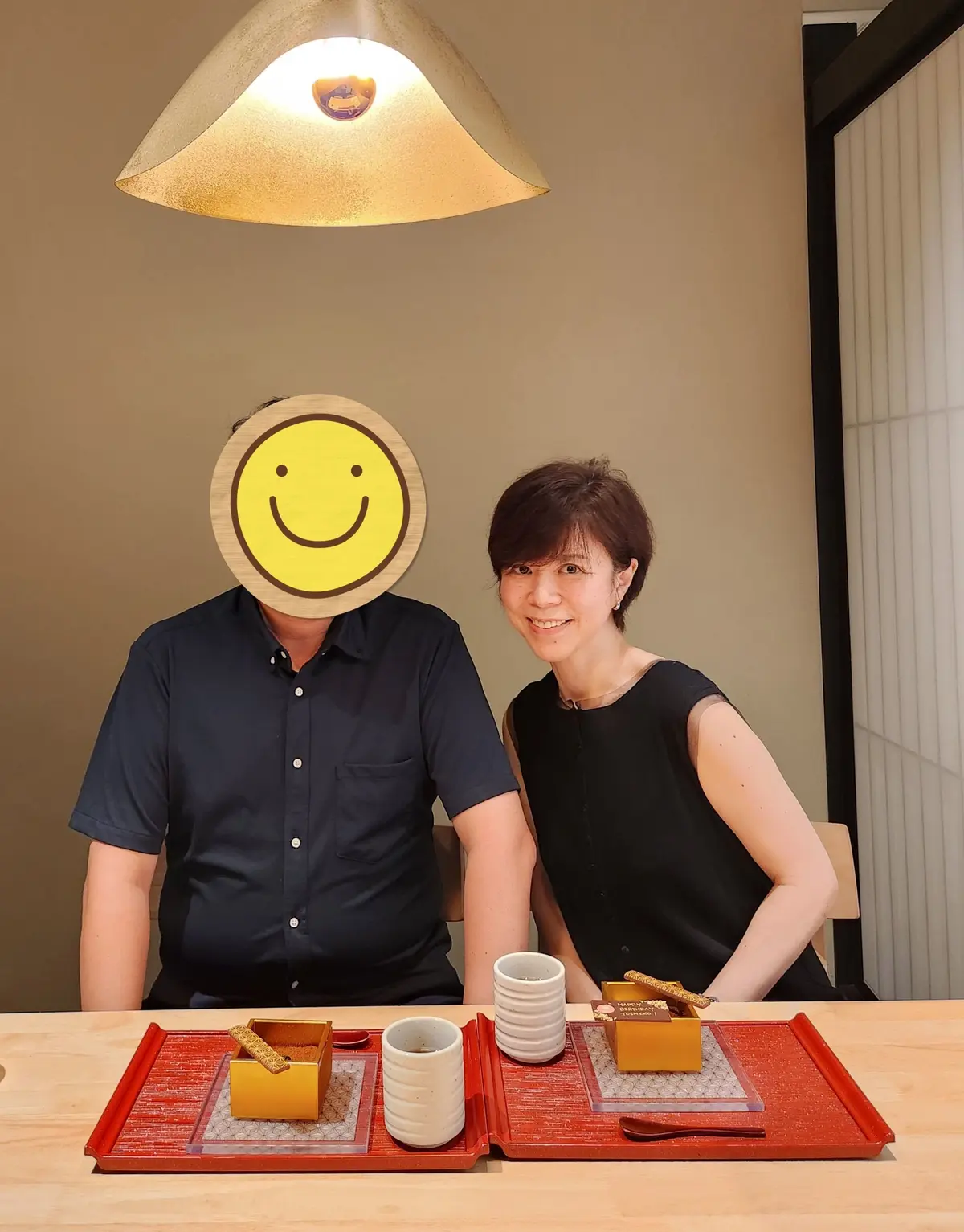 笑顔の男女が、木製のテーブルに置かれたお菓子と飲み物を囲んで写っている。男性は顔の一部が黄色いイラストで隠れており、黒いシャツを着ている。女性は黒いワンピースを着て微笑んでいる。テーブルの上には、赤いトレイにお菓子が入った箱と白いカップが並んでいる。