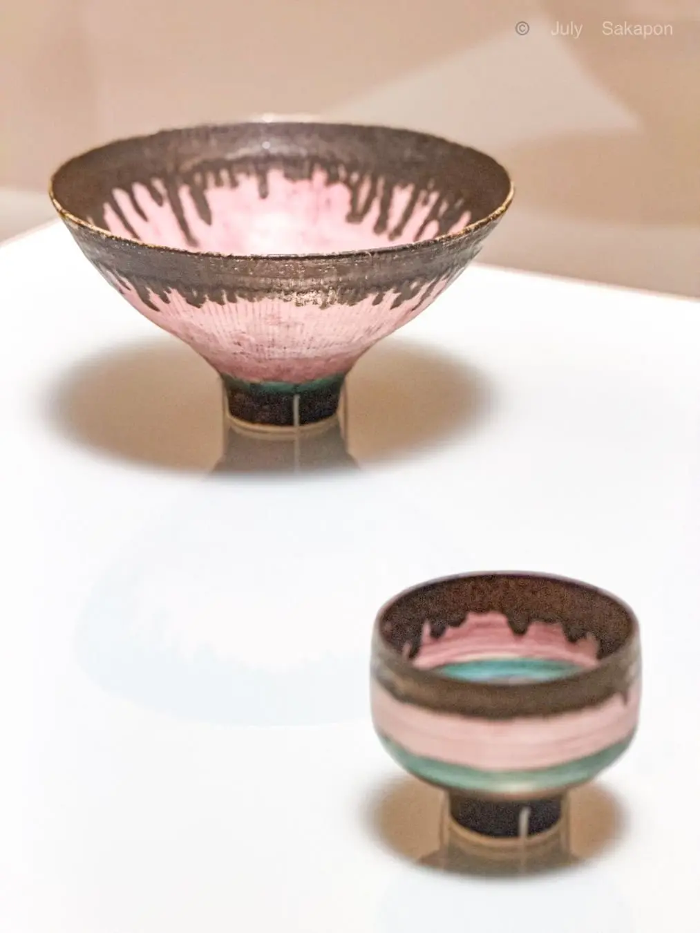 ピンクと茶色の釉薬が施された、2つの陶器の器が白い台の上に置かれている。上部は浅いボウル型、下部は細長いグラス型。それぞれに黒い装飾が施され、光沢のある質感。