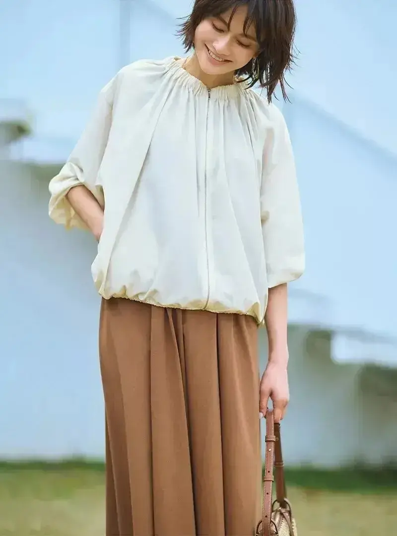 ベージュのプルオーバーと茶色のロングスカートを着た女性が、笑顔で草原に立っている。右手に編みかご風の鞄を持っている。