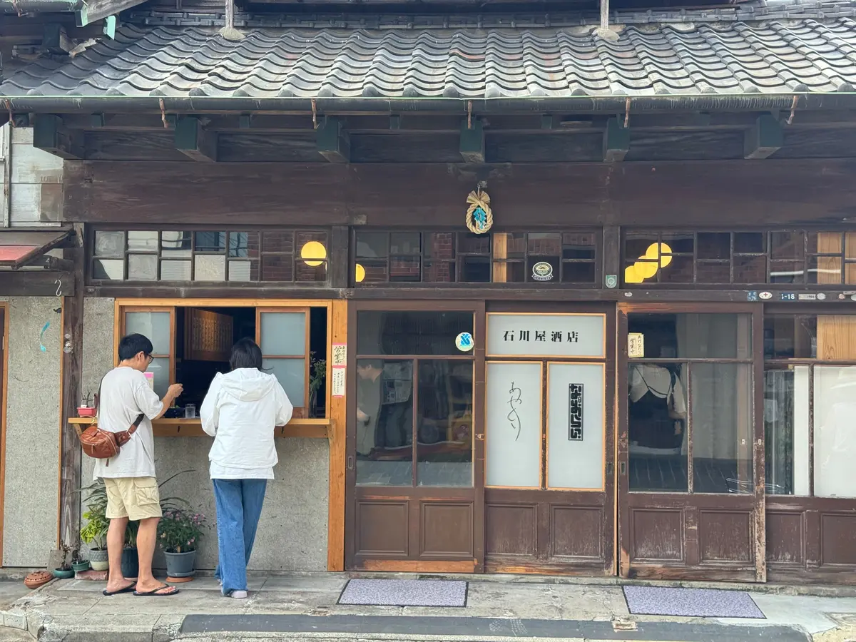 木造の建物に面した窓口で、2人が商品を選んでいる様子。窓口には漢字で「石川屋酒店」と書かれた看板がある。建物の外観は古風で、緑色の装飾が施されている。
