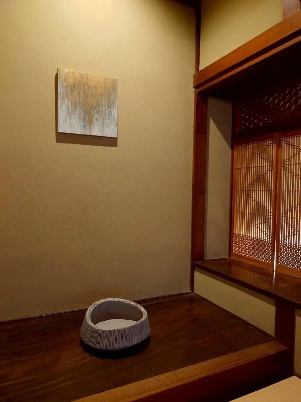 茶色の壁に絵画が飾られ、床には編み込み模様の器が置かれている。右側には木製の窓枠と、格子状の障子が見える。全体的に落ち着いた色調で、和風の空間を表現している。