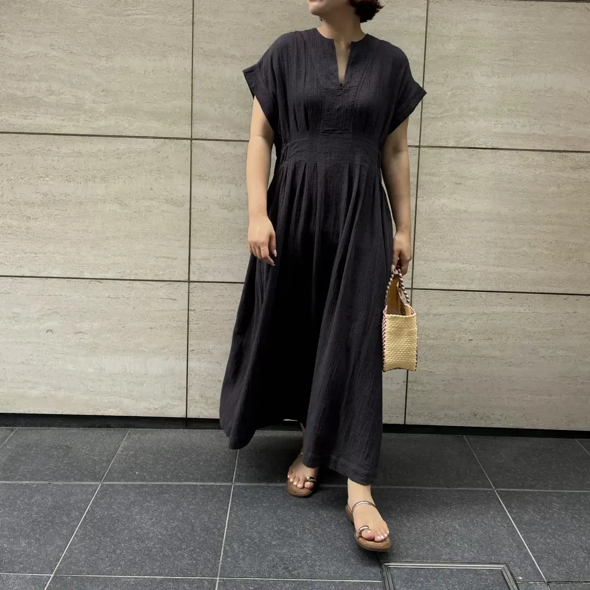 黒いギャザーワンピースを着た女性が立っている写真。ひざ下丈で、ゆったりとしたシルエット。茶色の編み込みバッグを持ち、足元はシルバーのサンダル。背景はベージュのタイル壁。
