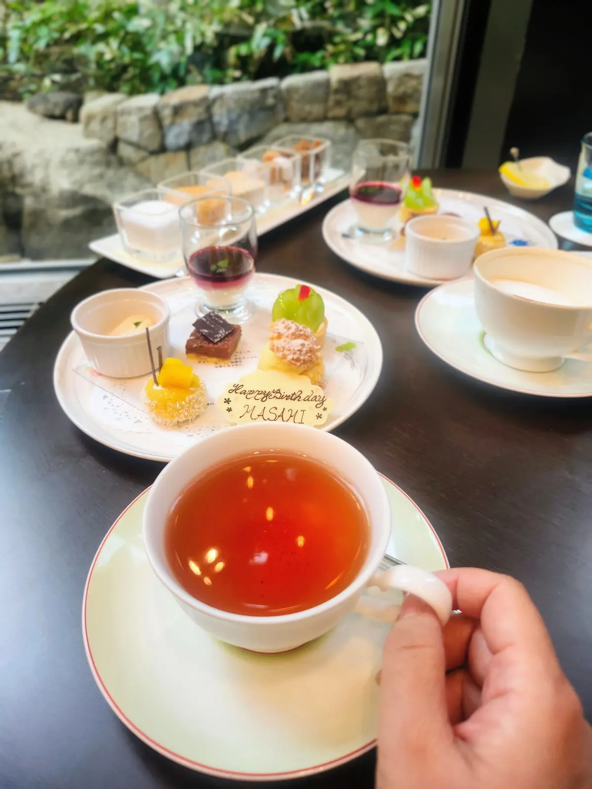 テーブルに並んだ誕生日のお祝いのデザートと飲み物。茶色の紅茶、グラスに入ったゼリーやケーキ、サンドイッチなどが盛り付けられた白いお皿が並び、背景には緑の植物が見える。