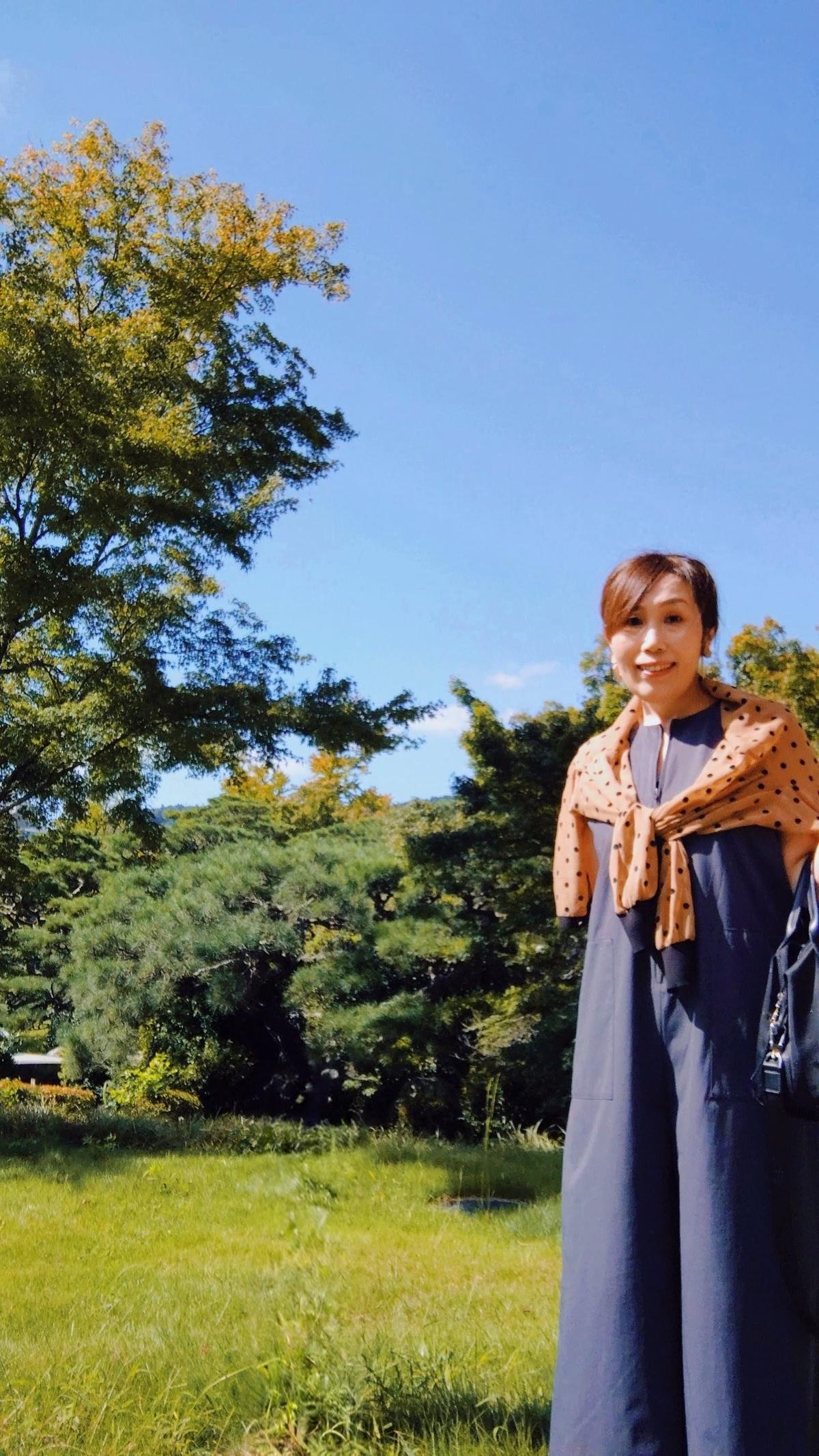 青空の下、緑の芝生に立つ女性。濃い青色のつなぎにオレンジ色のドット柄のスカーフを羽織り、笑顔を見せている。背負い鞄を背負っている。背景には緑豊かな木々が見える。