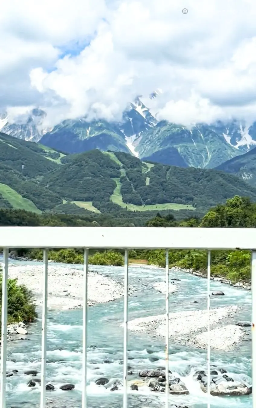 山々と川の風景、バルコニーの手すりから見た景色、雪をかぶった山々、緑の森、岩が散在する川