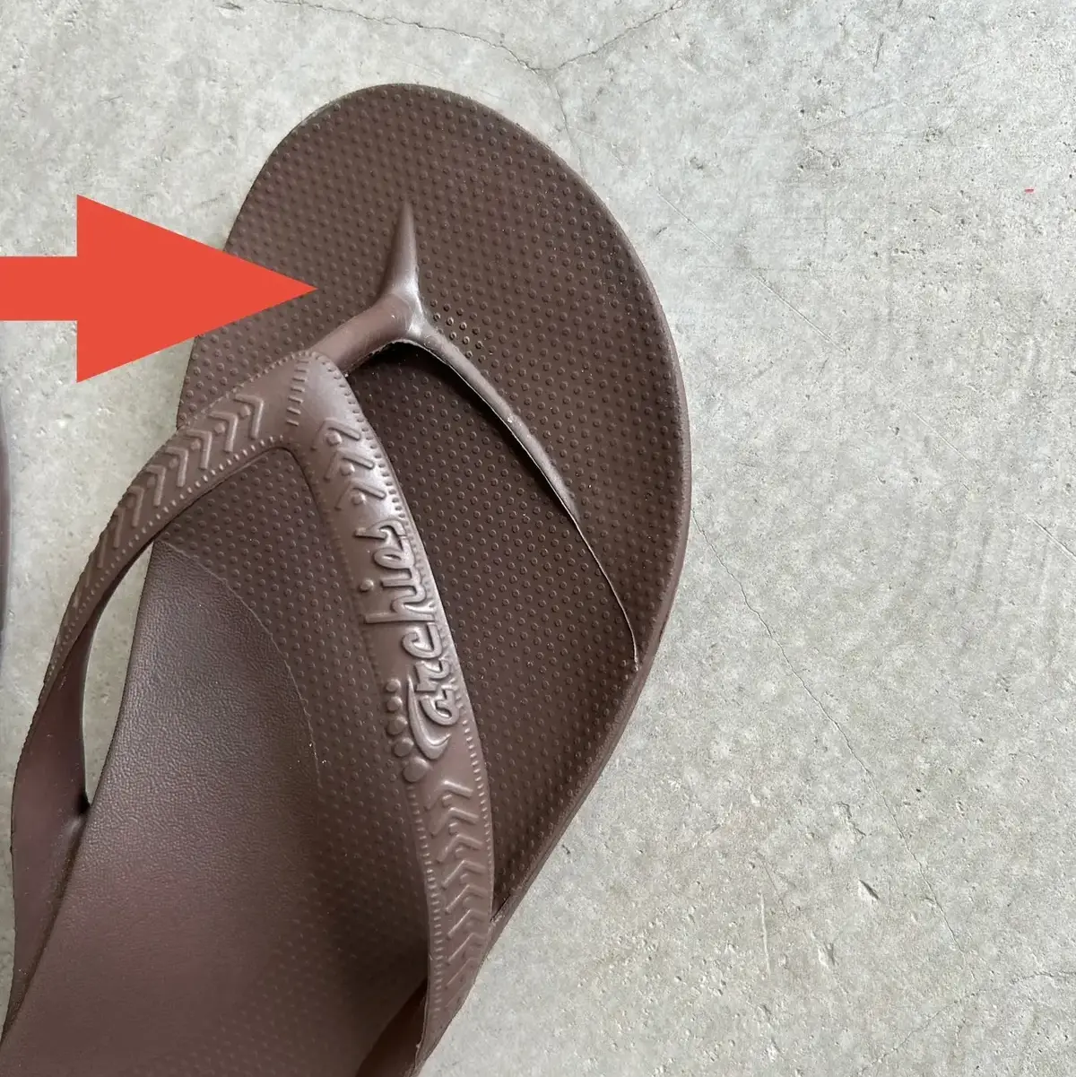 茶色のフリップフロップが地面に置かれている。左端には小さな穴が開いている。ソールには「Aloha Soles」のロゴが刻印されている。