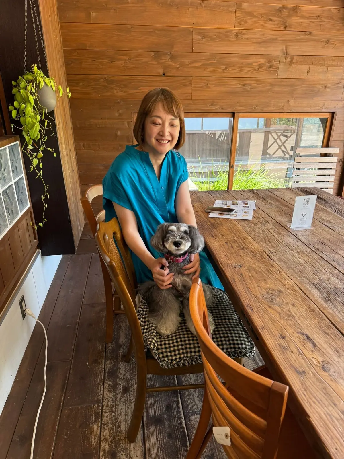 笑顔の女性が、膝の上に座った灰色の小型犬を優しく抱いている。木製のテーブルと椅子が置かれた、木材を多用した明るい空間。