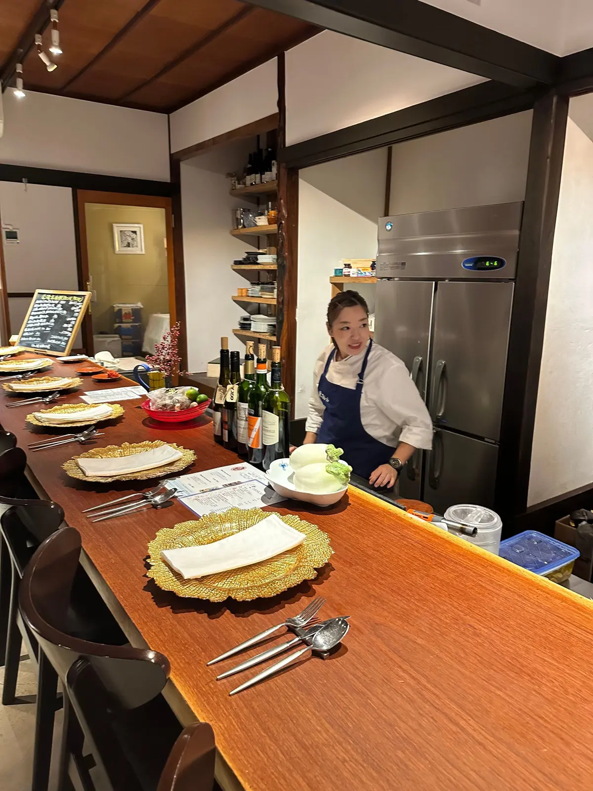 木製の長テーブルに並べられた食器とカトラリー。中央には白いテーブルクロスとメニューが置かれ、背景には厨房でエプロン姿の女性が立っている。棚には食器が並び、冷蔵庫がある。