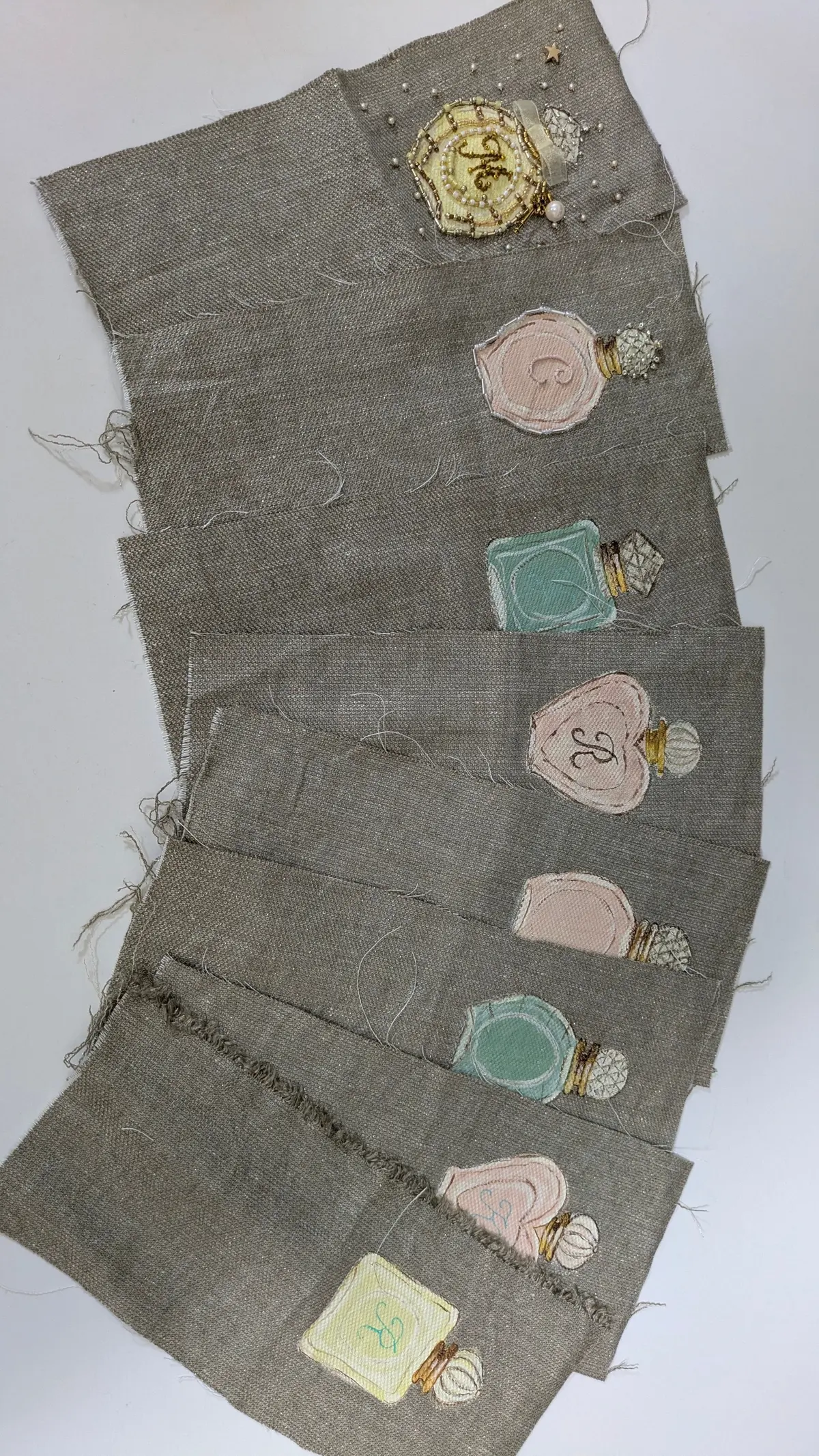 麻布地に、刺繍とビーズで装飾された香水瓶のモチーフが並ぶ。香水瓶の色は、ピンク、水色、ベージュなど様々で、それぞれ異なるデザイン。手作りの温かみのある作品。