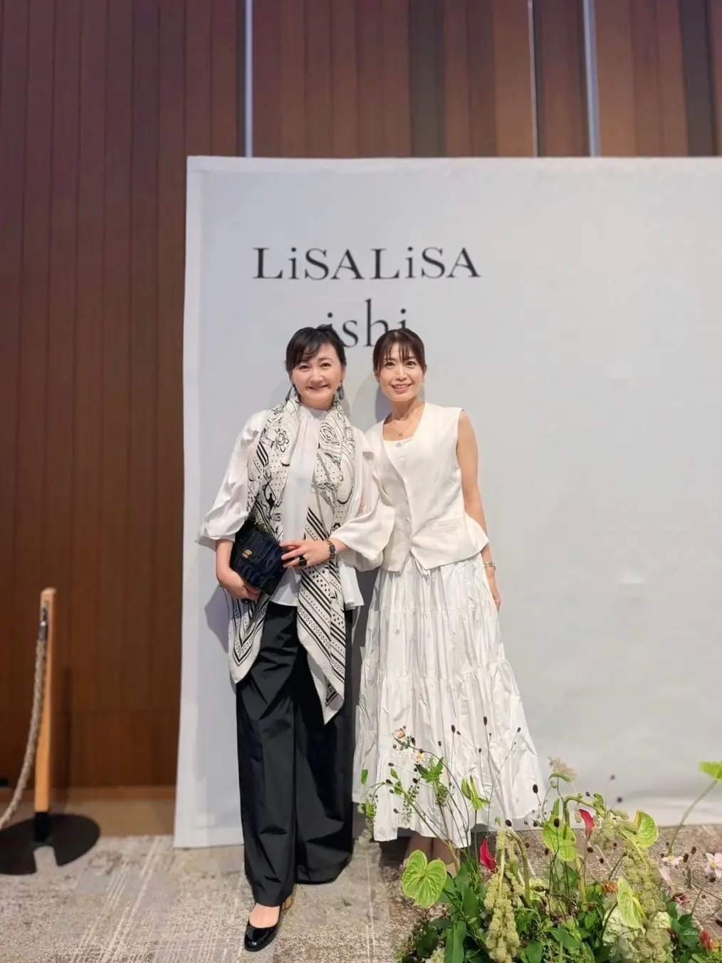 LiSA LiSAのロゴを背景に、2人の女性が並んで立っている写真。左の女性は黒いパンツスーツに白いスカーフ、右の女性は白いタンクトップにフリル付きのスカートを着用。それぞれ笑顔でこちらを見ている。
