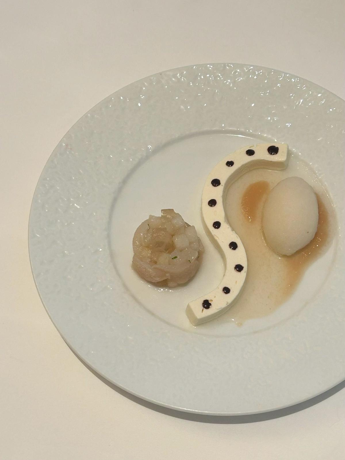白い器に盛り付けられた、上品な料理。真ん中に白身魚、左に刻み野菜と魚の和え物、右には horseshoe 型の白い飾りと黒い点々が配置されている。
