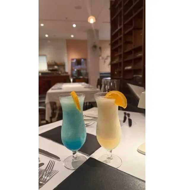 テーブルに置かれた2つのカクテル。左は青色のフローズンカクテル、右は淡い黄色いフローズンカクテル。どちらもグラスの縁にオレンジのスライスが飾られている。背景にはレストランのテーブル席が見える。