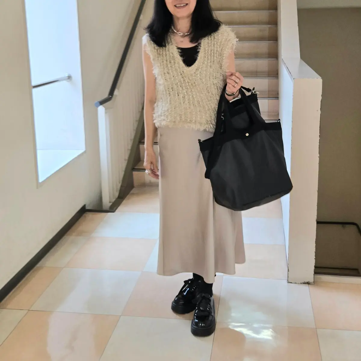 ベージュのスカート、白いニットベスト、黒いブーツを着用した女性が、階段の横でポーズをとっている。黒いトートバッグを持ち、ネックレスを身につけている。背景は明るいベージュ色の壁と階段。