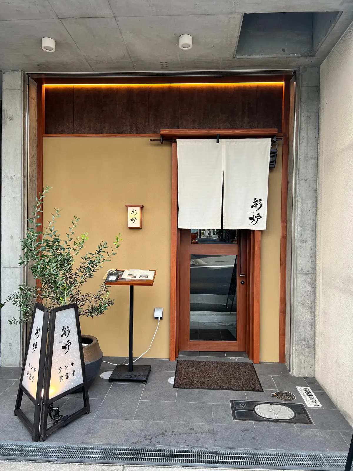 コンクリートの壁と木製のフレームで囲まれた入り口。右には白いのれん幕が、入り口の周りには看板が設置されている。看板には黒い文字で「彩炉」と書かれている。