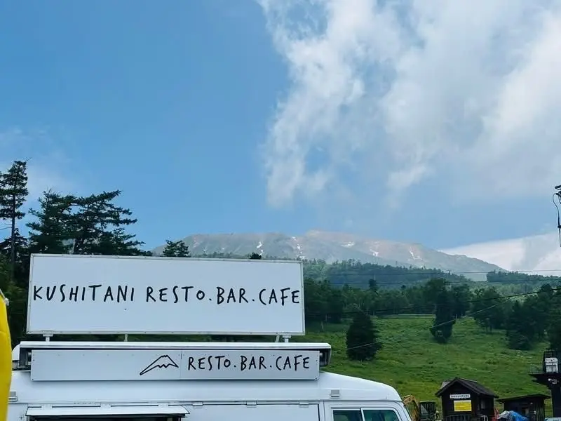 白のバンに「KUSHITANI RESTO.BAR.CAFE」のロゴと山型のイラストが描かれ、背景には雪を抱いた山々が広がる風景。