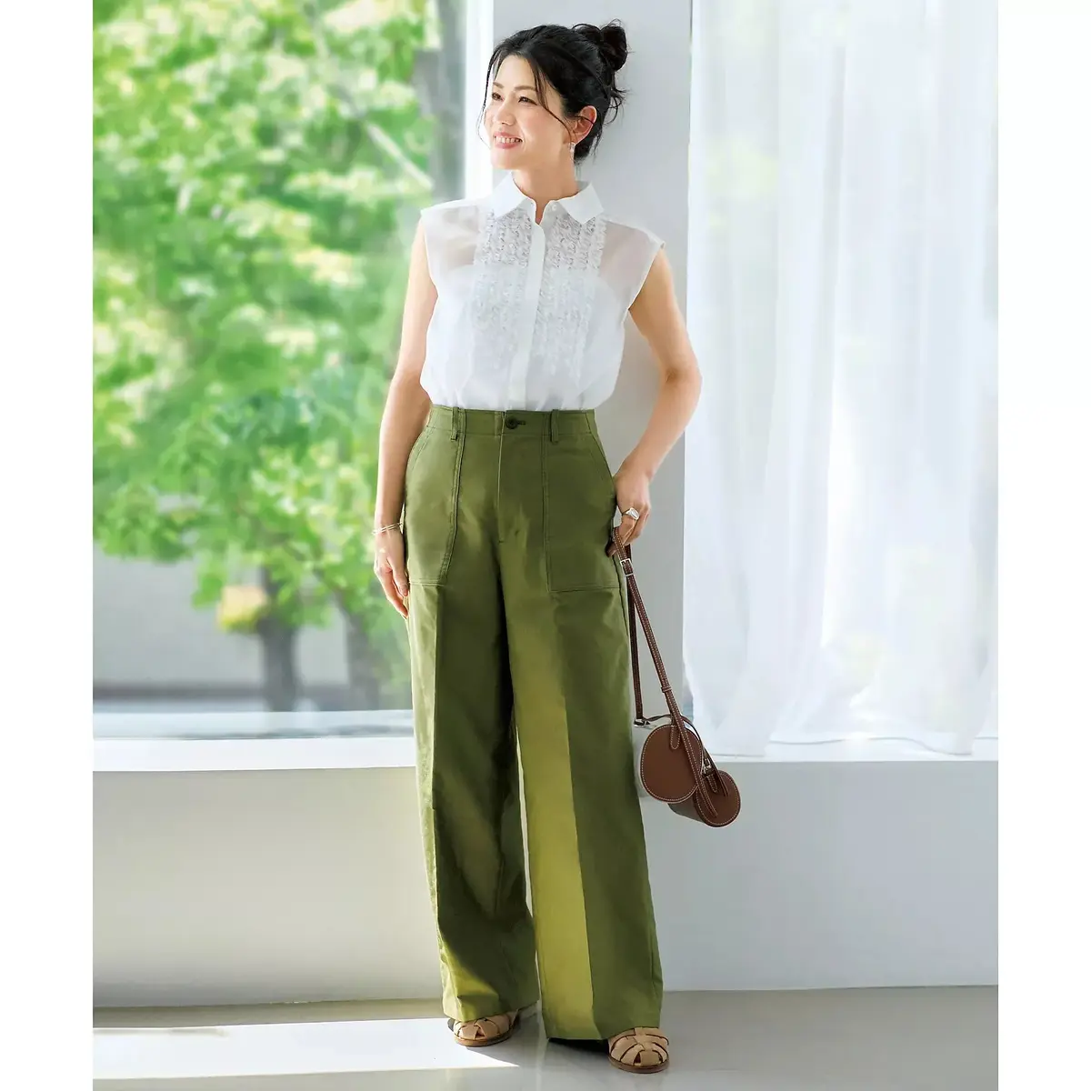 白のフリルシャツに、深緑色のワイドパンツを合わせた女性。窓際で微笑み、茶色のショルダーバッグを持つ。まとめ髪で、ゴールドのアクセサリーを身に着けている。