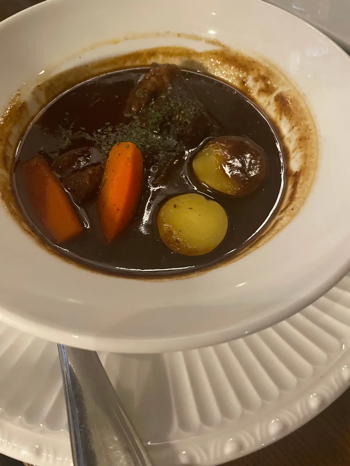 白い皿に盛り付けられた、牛肉と野菜の煮込み料理。牛肉、人参、ジャガイモが、濃い茶色のソースに浸かっている。皿の縁にはソースが飛び散り、仕上げに緑色のハーブが散らされている。