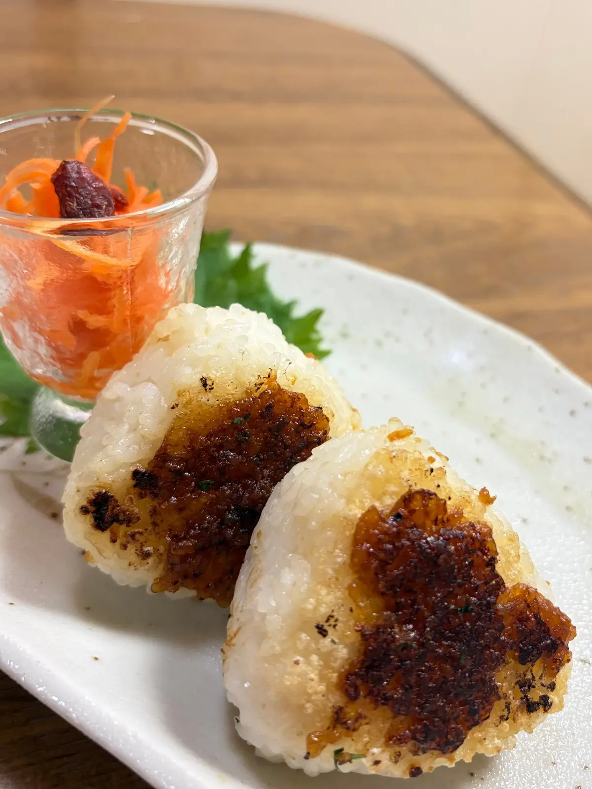 白ご飯に甘辛く照り焼きにした豚肉が乗ったおにぎり2個と、千切りキャロットの漬物がガラスの器に入った盛り合わせ。白い皿に、緑の葉を添えて置かれている。