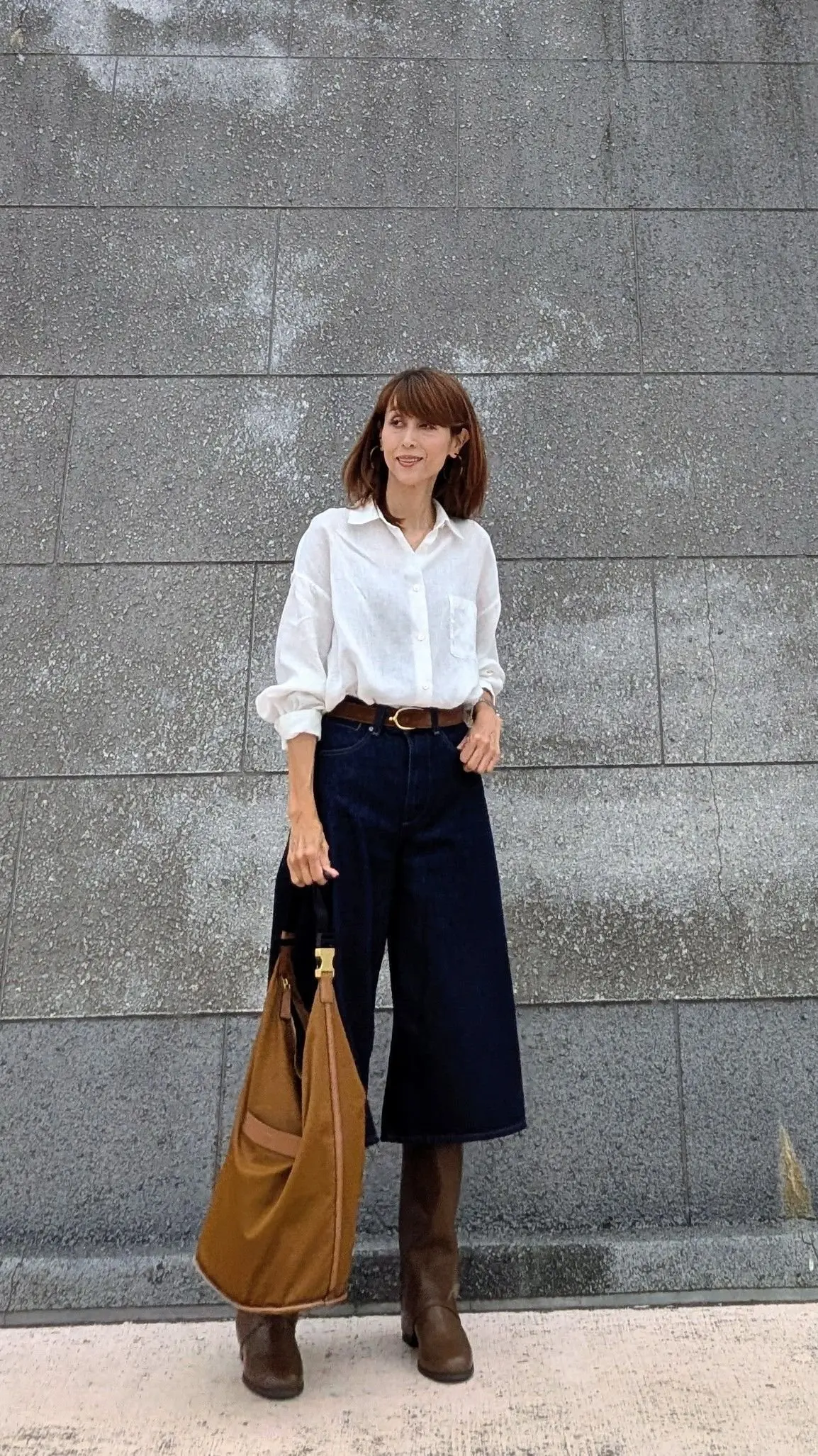 白いボタンダウンシャツに濃い青のワイドパンツ、茶色のロングブーツを合わせた女性。茶色のトートバッグを持ち、グレーの壁の前で立っている。