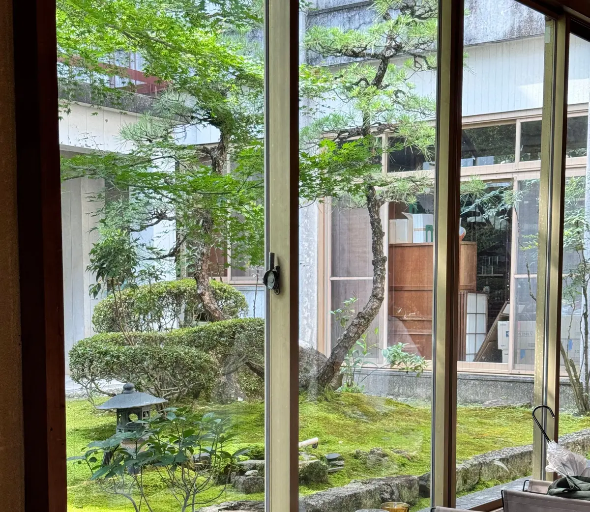 窓から見える日本庭園と室内テーブル

窓ガラス越しに緑豊かな日本庭園が広がり、芝生や岩、盆栽が配置されている。室内には丸いテーブルにグラスやプレートが置かれ、モダンな椅子が配置されている。
