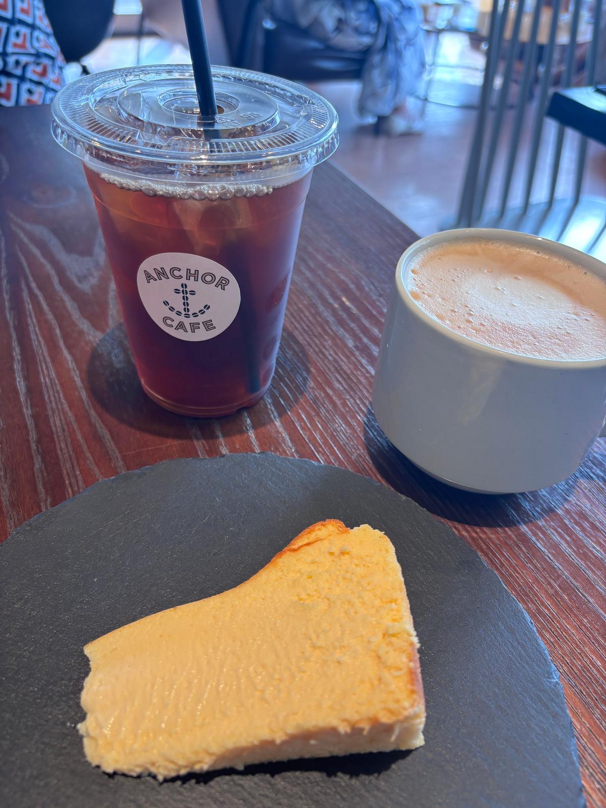 Anchor Cafeのロゴが入ったアイスコーヒー、ミルクを泡立てたコーヒー、チーズケーキが黒いお皿に置かれている。