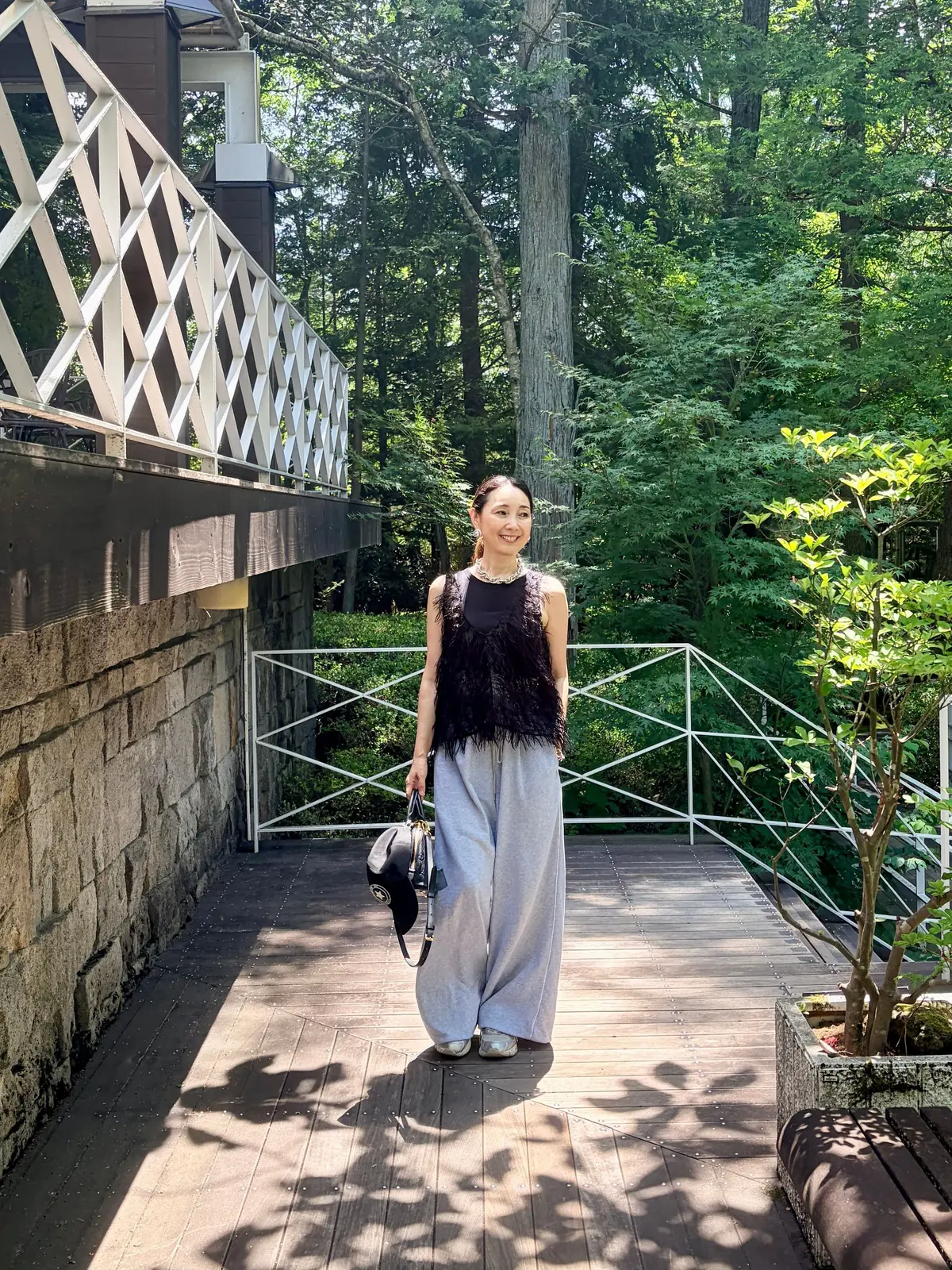 木陰の遊歩道に立つ女性。黒の羽根飾りのトップスにグレーのワイドパンツ、シルバーの靴を着用。右手に黒いバッグを持ち、微笑んでいる。背景には緑豊かな木々と橋が見える。