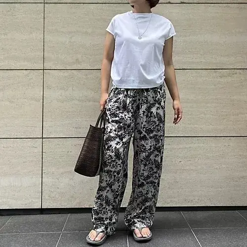 白いTシャツに、黒と白の植物柄のワイドパンツを合わせた女性。茶色のバッグを持ち、白いサンダルを履いている。背景はベージュ色のタイル壁。