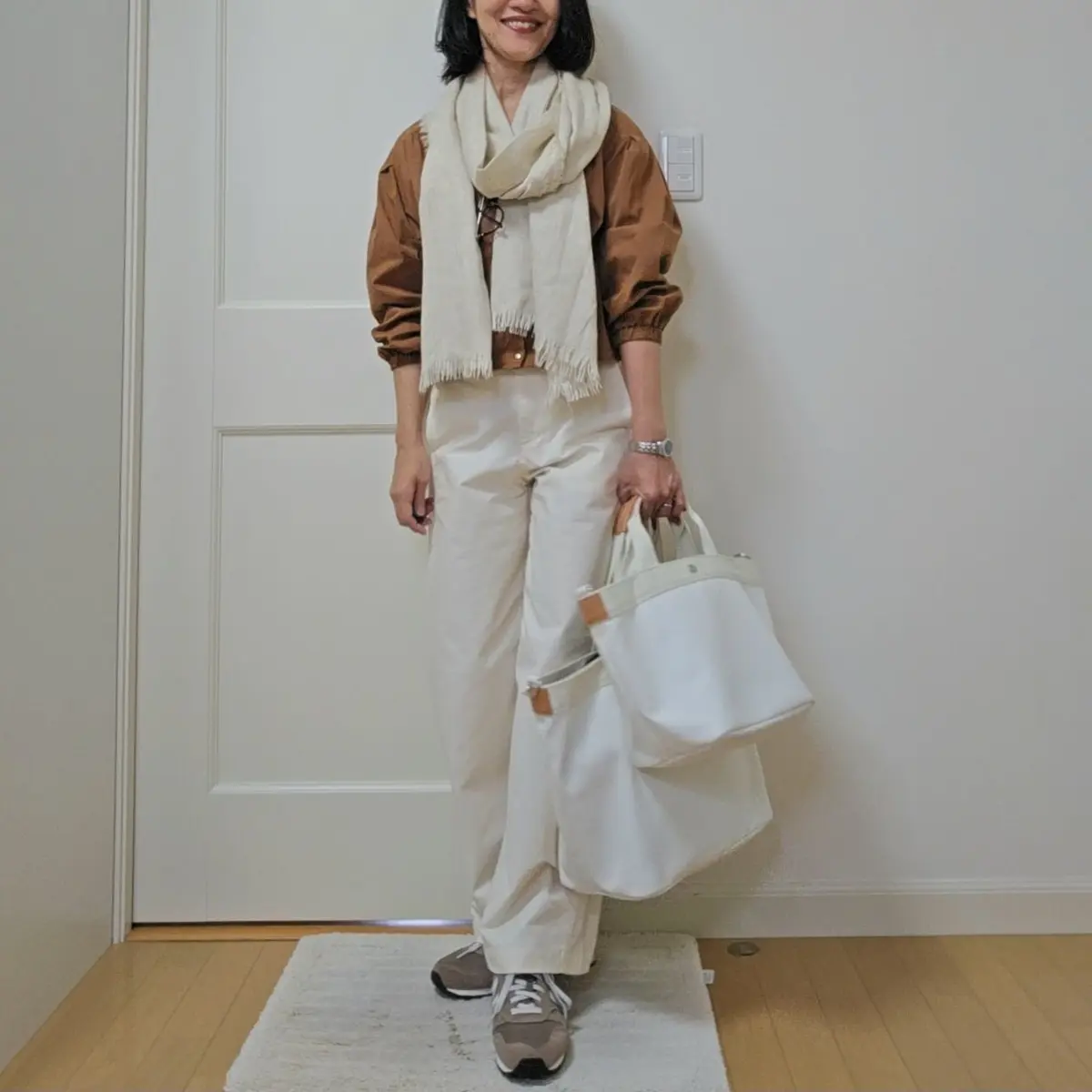 ブラウンのトップスに白いパンツ、ベージュのストールを巻いた女性。白いトートバッグを手に持ち、グレーのスニーカーを履いている。背景は白い壁と木製の床。