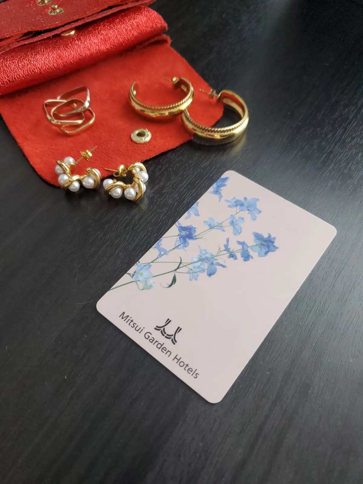 赤い革の布の上に金色のイヤリングやリングが散らばり、青い花が描かれた「Mitsui Garden Hotels」のカードが置かれている
