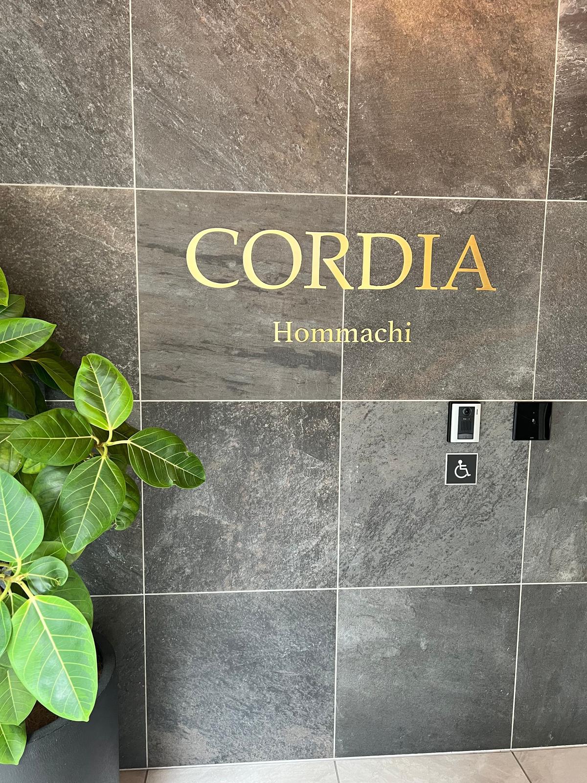 石張りの壁に設置された「CORDIA Hommachi」の銘板。銘板の下には、点字ブロックと車椅子マークの表示がある。左側には緑の葉の植物が置かれている。