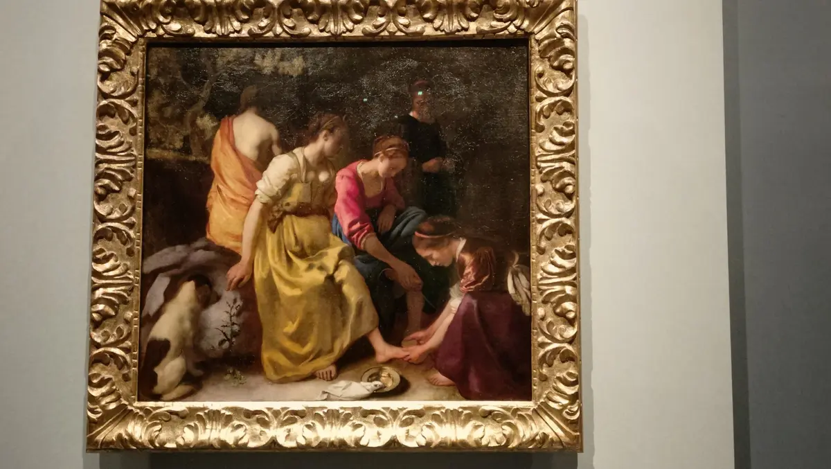 金色の装飾的な額縁に入った絵画。画面中央には、水辺で女性が足湯をしている場面が描かれている。左には観る女性、右には見守る男性、背景には木々が茂り、遠くに人物が見える。全体的に落ち着いた色調で、古典的な雰囲気を醸し出している。