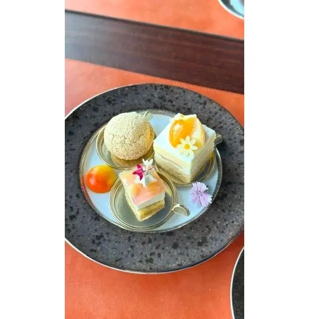 黒とグレーの模様の皿に、3種類のミニケーキと、赤と黄色のミニトマトが置かれている。それぞれのケーキは小さなスプーンに乗せられ、食用花やフルーツで飾られている。