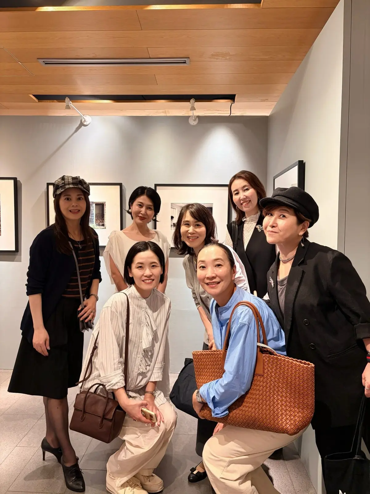 美術館のような空間で、7人の女性が笑顔で写っている集合写真。黒のジャケットや帽子を被る人もおり、それぞれ異なるファッションを楽しんでいる。背景には白黒の絵画が飾られている。