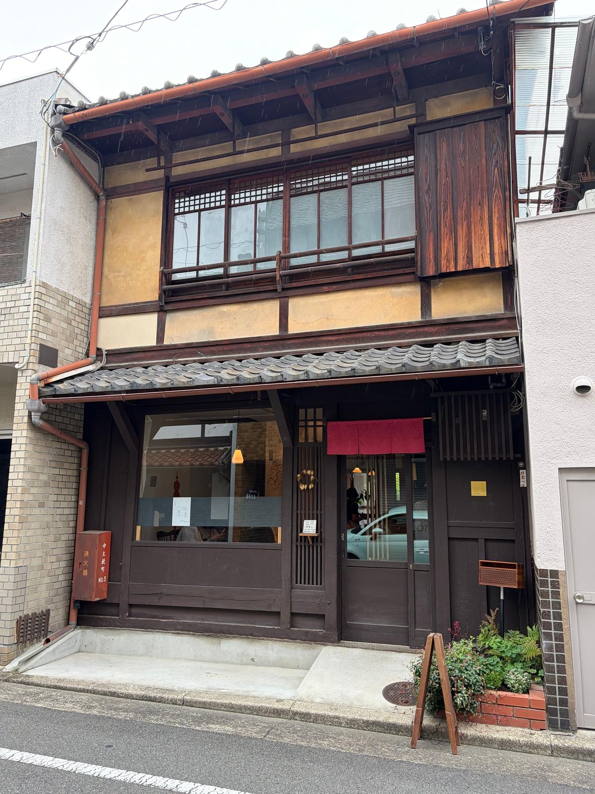 伝統的な二階建ての木造建築。茶色い壁と赤い屋根、木製のバルコニーと窓が特徴。入口には赤い提灯とガラスのドアがあり、店内が見える。左側に小さな看板と植えられた植物がある。
