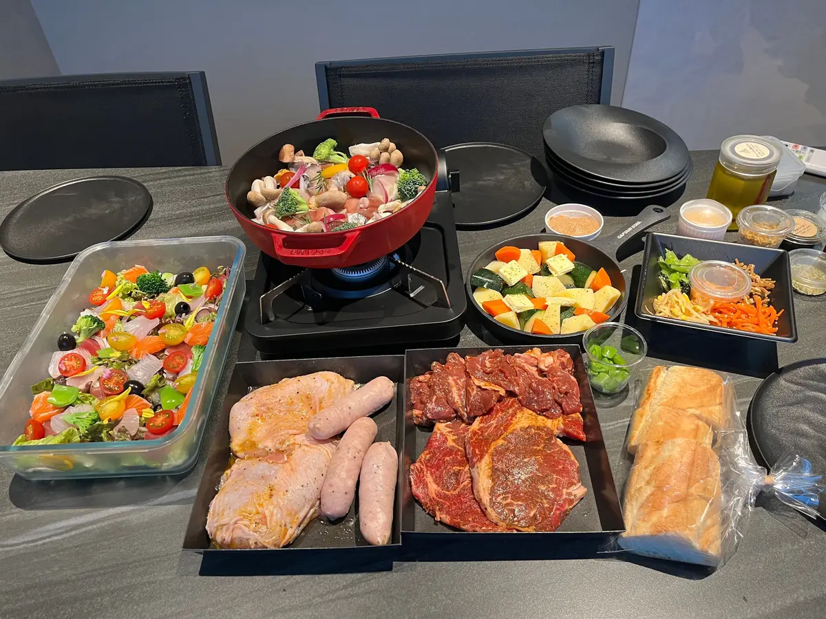 テーブルに並んだ食材。赤色のスキレットで野菜炒め、サラダ、鶏肉、ソーセージ、牛肉、野菜の炒め物、パン、調味料などが置かれている。ポータブルガスコンロがスキレットの下にある。