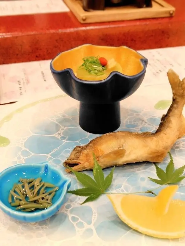 青い台に、焼き魚とレモン、豆、緑の葉、小さな青い器に具材が乗った料理が並ぶ