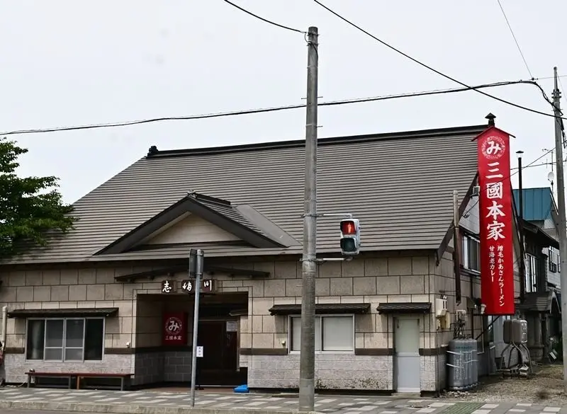 屋根付きの建物の外観。茶色のタイル屋根とベージュ色の壁。看板には赤い文字で「三國本家」と書かれている。道路を挟んで隣には別の建物がある。